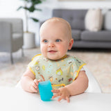 A smiling baby wearing a green bib in a high chair holds a Bumkins Silicone Starter Cup: Blue.