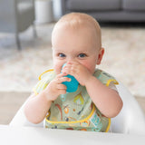 A baby in a high chair joyfully sips from a Bumkins Silicone Starter Cup: Blue, wearing a camping-themed bib.