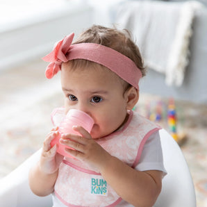 A baby wearing a pink headband and bib in a high chair drinks from a Bumkins Silicone Starter Cup: Pink.