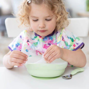 A curly-haired child in a colorful bib sits at a white table, removing a Bumkins Silicone Stretch Lid for Grip Bowl from a green bowl.