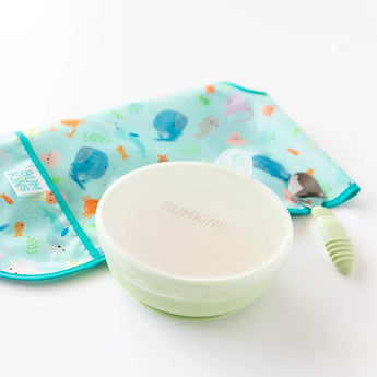 Bumkins Silicone Stretch Lid for Grip Bowl covers a green bowl next to a green spoon and a folded ocean-themed bib on a white background.
