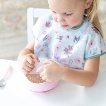 A young girl in a floral bib removes a Bumkins Silicone Stretch Lid for Grip Bowl  from a pink bowl of cereal and milk at a white table.