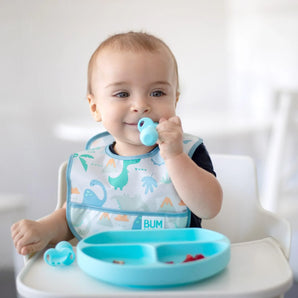 A baby in a high chair wears a Bumkins Starter Bib 2 Pack: Dinosaurs & Blue Tropic, eating from a blue divided plate with blue baby utensils.