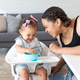 A woman smiles at a toddler in a high chair wearing a Bumkins Starter Bib 2 Pack: Floral & Lace with a blue bowl on the tray.
