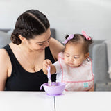 A woman smiles as baby, in a pink Bumkins Starter Bib 2 Pack: Floral & Lace, reaches for a purple bowl on a white table.