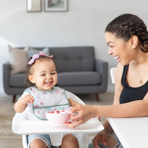A smiling mom feeds a happy toddler in a high chair from a pink bowl; the toddler wears a Bumkins Starter Bib 2 Pack: Floral & Lace.
