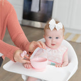 A baby in high chair with white bow wears a Bumkins Starter Bib 2 Pack: Floral & Lace as an adult places a pink bowl on the tray.