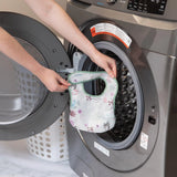 A person holds a Bumkins Starter Bib 2 Pack: Floral & Lace in front of an open front-loading washing machine.