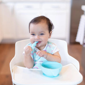 A smiling baby in a high chair holds a spoon, wearing a Bumkins Starter Bib 2 Pack: Rainbows & Unicorns with a blue bowl on the tray.