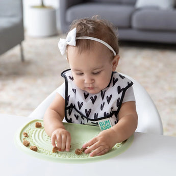 A baby in a high chair in a black & white bib plays with cereal pieces on a placemat from Bumkins Baby-Led Weaning Deluxe 7-Piece Set: Sage.