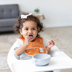 A toddler with curly hair wears an orange Bumkins Starter Bib 2 Pack: Sunshine and Grounded, eating with a spoon in a high chair.