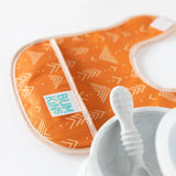 Close-up of an orange Bumkins Starter Bib 2 Pack: Sunshine and Grounded beside a gray baby bowl and spoon on a white background.