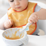 A baby wearing a yellow Bumkins Starter Bib 2 Pack: Sunshine and Grounded in a high chair with a spoon, eating from a bowl of mushy food.