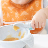 Close-up of a child in an orange Bumkins Starter Bib 2 Pack: Sunshine and Grounded, holding a spoon of baby food over a bowl.