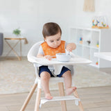 In a bright room, a toddler in a high chair uses a spoon to eat while wearing an orange Bumkins Starter Bib 2 Pack: Sunshine and Grounded.