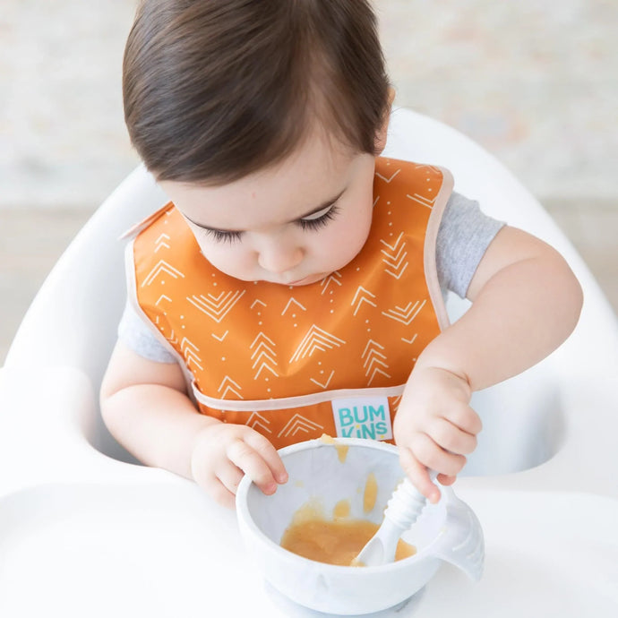 A toddler in a high chair eats from a gray bowl with a spoon while wearing an orange Bumkins Starter Bib 2 Pack: Sunshine and Grounded.