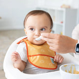 An adult feeds a baby with a spoon while they sit in a high chair, wearing a yellow Bumkins Starter Bib 2 Pack: Sunshine and Grounded.
