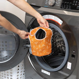 A person holds an orange Bumkins Starter Bib 2 Pack: Sunshine and Grounded in front of an open front-loading washing machine.