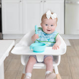 A baby sits in a high chair, wearing a Bumkins Starter Bib 2 Pack: Ocean Life & Whale Tail and holding a blue spoon over a blue bowl.