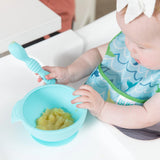 A baby in a Bumkins Starter Bib 2 Pack: Ocean Life & Whale Tail holds a spoon over a bowl of mashed food on a high chair tray.