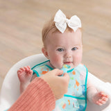 A baby in a Bumkins Starter Bib 2 Pack: Ocean Life & Whale Tail sits in a high chair, being spoon-fed by an adult hand.
