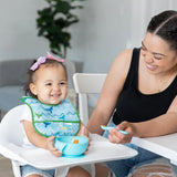 A smiling woman feeds a happy toddler in a high chair wearing a Bumkins Starter Bib 2 Pack: Ocean Life & Whale Tail from a blue bowl.