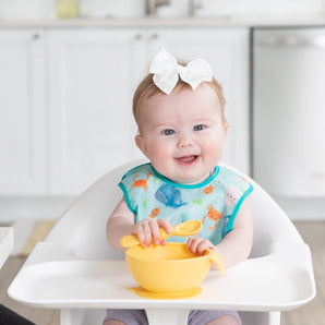 A baby in a high hair smiles, wearing a Bumkins Starter Bib 2 Pack: Ocean Life & Whale Tail while holding a yellow bowl and spoon.