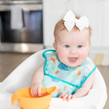 A baby in a bow wears a Bumkins Starter Bib 2 Pack: Ocean Life & Whale Tail, sitting in a high chair and holding an orange bowl and spoon.