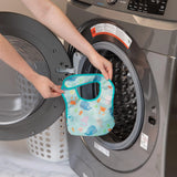 A person holds a Bumkins Starter Bib 2 Pack: Ocean Life & Whale Tail in front of an open front-loading washing machine.