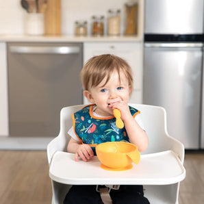 A toddler in a high chair, wearing a Bumkins Starter Bib 2 Pack: Jungle & Animal Prints, chews a spoon with an orange bowl nearby.