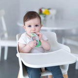 A baby in a high chair chews on a Bumkins Silicone Teething Charms: Green, with a blurred table and flowers in the background.