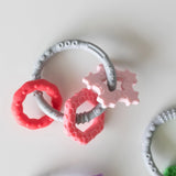 Bumkins Silicone Teething Charms: Pink rests on a white background with another teething toy nearby.