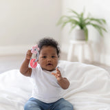 A baby in a white shirt and jeans sits on a white blanket, holding a Bumkins Silicone Teething Charms: Pink, with a potted plant behind.