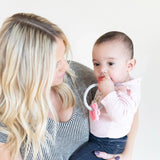 A woman with long blonde hair holds a baby in a pink shirt chewing on a Bumkins Silicone Teething Charms: Pink.