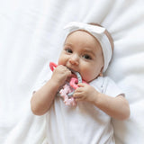 A baby in white outfit with a headband chews a Bumkins Silicone Teething Charms: Pink while laying on a white bedspread.