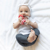 A baby wearing a bow headband on a white blanket, smiling softly, chews a Bumkins Silicone Teething Charms: Pink.