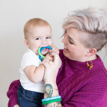 A smiling woman with short blonde hair wearing a purple sweater holds a baby chewing on Bumkins Silicone Teething Rings 4 Pack: Summer.