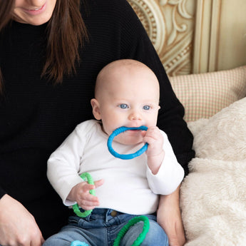 A baby in a white shirt holds and chews Bumkins Silicone Teething Rings 4 Pack: Summer, sitting on an adult's lap in a cozy room.