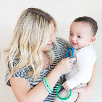 A woman smiles at a baby, wearing Bumkins Silicone Teething Rings 4 Pack: Summer while holding another.