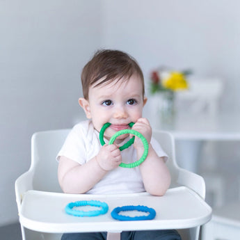 A baby in a high chair chews on Bumkins Silicone Teething Rings 4 Pack: Summer in a bright room with flowers on a table in the background.