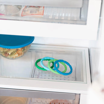 Bumkins Silicone Teething Rings 4 Pack: Summer sits on a glass refrigerator shelf, with a covered dish of food nearby.