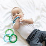 A baby on a white blanket, wearing a onesie and pants, chews a Bumkins Silicone Teething Rings 4 Pack: Summer while the others lay nearby.