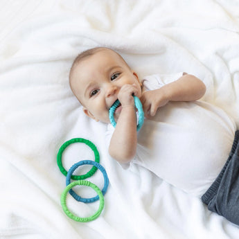 A baby on a white blanket chews a Bumkins Silicone Teething Rings 4 Pack: Summer, while the others sit nearby.