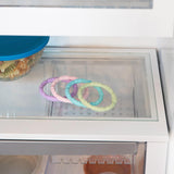 Bumkins Silicone Teething Rings 4 Pack: Spring sits beside a blue-lidded bowl on a glass refrigerator shelf.