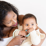 A smiling woman holds a baby, dressed in a beige shirt, chewing on a Bumkins Silicone Teething Rings 4 Pack: Spring.