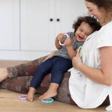 A woman sits with a laughing toddler holding Bumkins Silicone Teething Rings 4 Pack: Spring on her lap on a wood floor in a cozy home.
