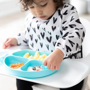 A toddler in a Bumkins Sleeved Bib: Hearts sits in a high chair, eating yogurt, fruit, & eggs from a blue divided plate.