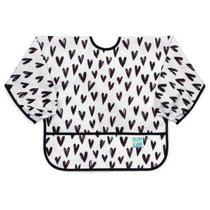 Bumkins Sleeved Bib: Hearts is a white waterproof bib with a black heart pattern, black trim, front pocket, and long sleeves.
