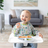 A happy toddler in a high chair enjoys spaghetti and fruit using a spoon while wearing a Bumkins Sleeved Bib: Cacti.