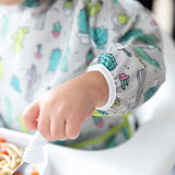 Close-up of a child in a high chair wearing a Bumkins Sleeved Bib: Cacti, holding a spoon.